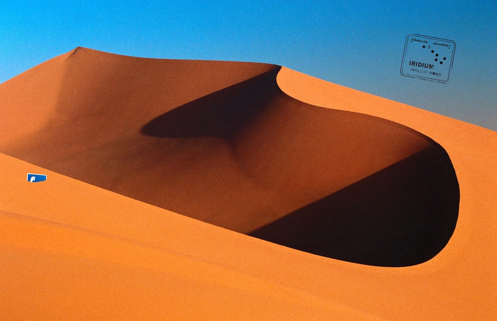 Iridium print ad: an entry stamp with Arabic script for Iridium Satellite Phones above an orange desert dune